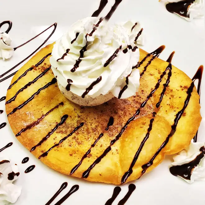 A golden brown pancake topped with vanilla ice cream, chocolate sauce, and whipped cream on a white plate.