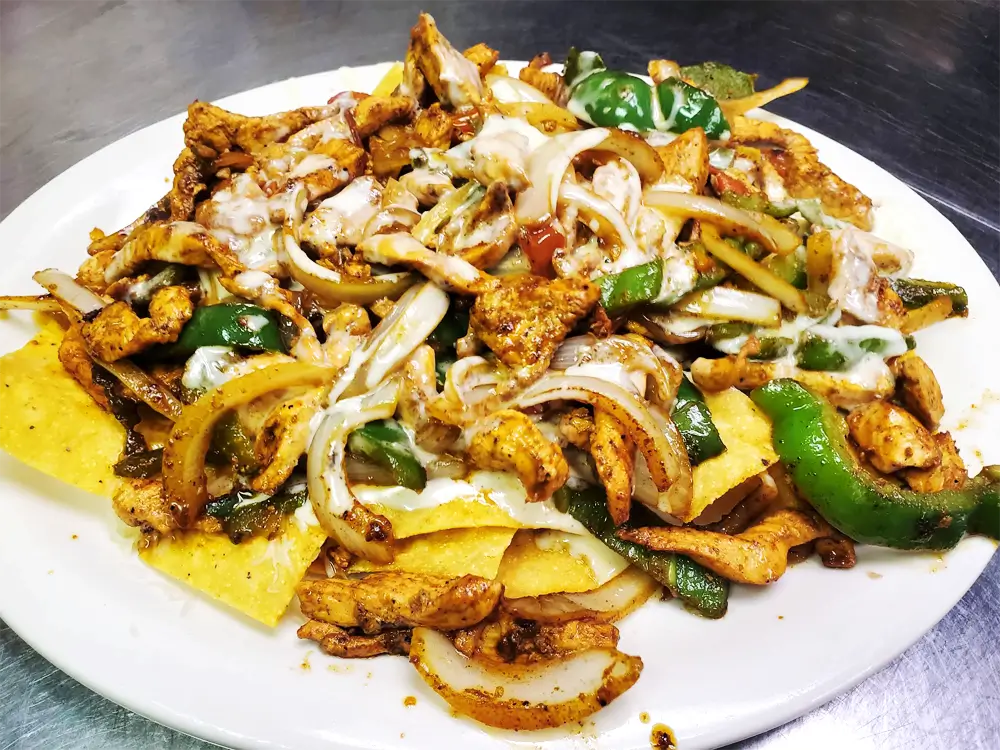 A white plate on a black surface holds a mix of brown chicken, green peppers, yellow onions, and white sauce, with crispy white chips.