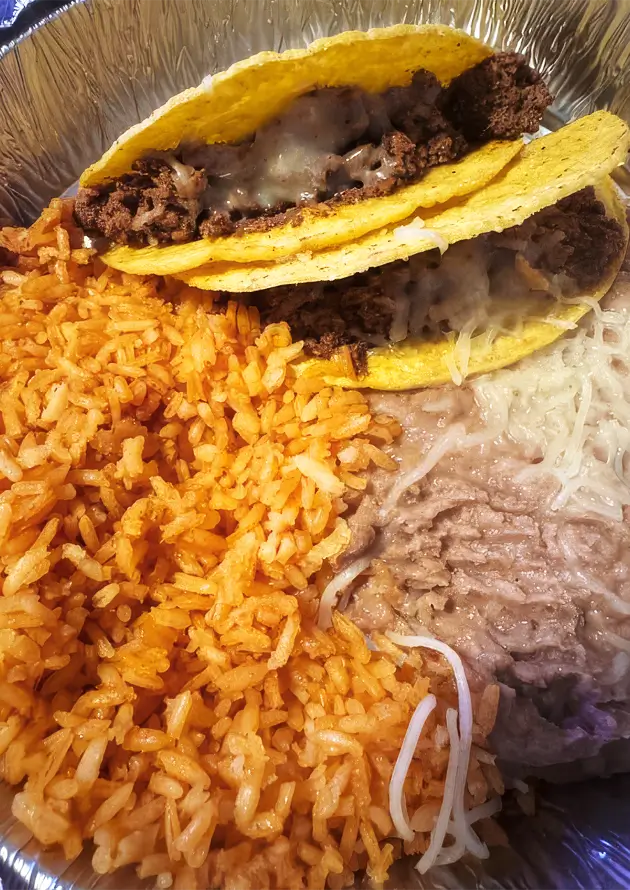 Colorful tacos served on a silver foil tray with orange rice, shredded cheese, and brown meat in a white plate and bowl.