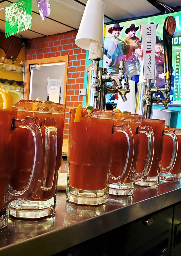 Metal bar counter with glasses of deep red drink, straws, and shiny metal finish, surrounded by signs and a beer machine.
