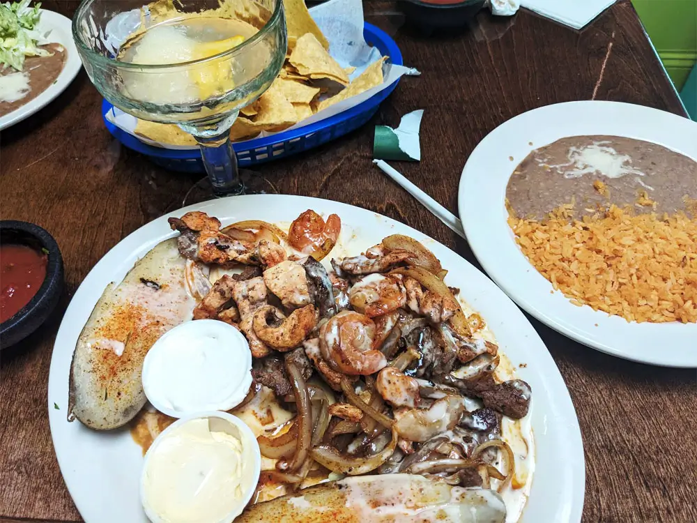 A wooden table set with a white plate of shrimp, tortillas, rice, water, salsa, and wine, against a green wall.